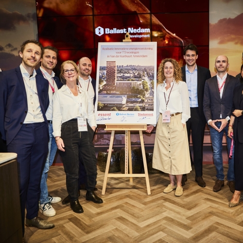 Groep aanwezigen poseert rond een presentatiestand met projectbord over de energievoorziening van Van der Kunbuurt in Amsterdam.