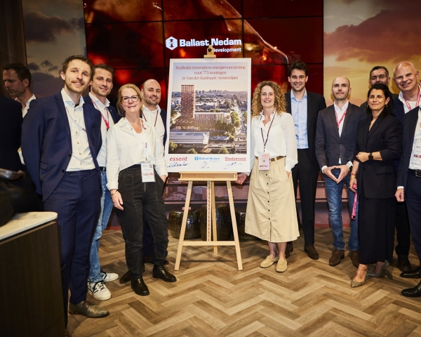 Groep aanwezigen poseert rond een presentatiestand met projectbord over de energievoorziening van Van der Kunbuurt in Amsterdam.